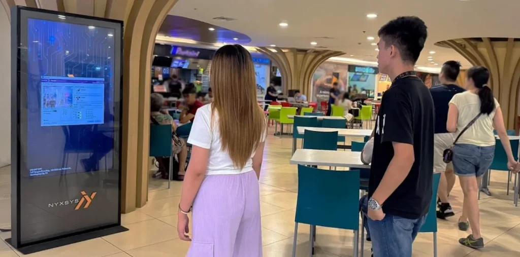 A vertical digital screen is shown on the left in a brightly lit indoor space, possibly a mall food court. The screen displays data and graphics, including a window with a graphical user interface. A woman with long brown hair, wearing a white shirt and light purple pants, is standing with her back to the camera, looking at the screen. A young man in a black t-shirt and blue jeans stands beside her, also looking at the screen. The bottom of the screen displays the NYXSYS logo. The background shows tables and chairs in the food court, with people seated and walking around.