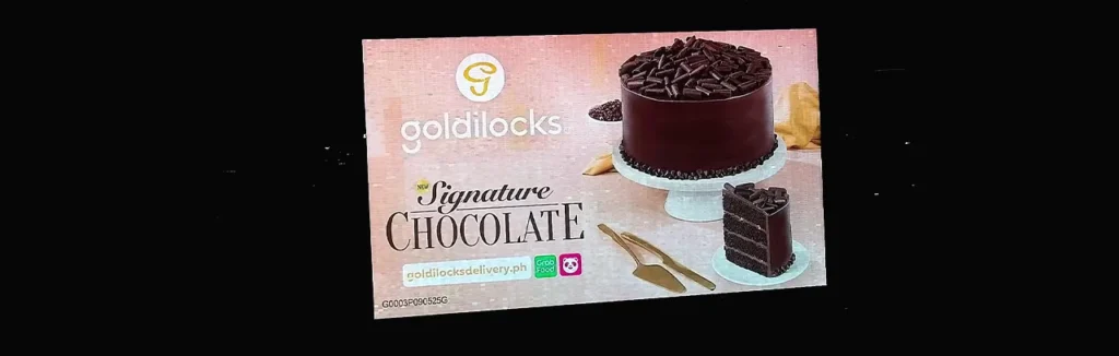 Digital billboard ad showing a full cake on a stand and a slice, with ordering options for goldilocksdelivery.ph and Grab Food.