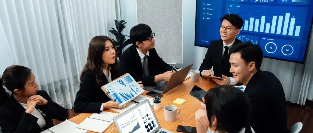A team of six professionals in black business suits and ties are gathered around a wooden conference table in a modern office setting. They are engaged in a discussion, looking at reports and digital screens. One woman is holding up a clipboard with charts and graphs. Laptops displaying data dashboards are open on the table, alongside coffee mugs and notebooks. In the background, a large digital screen displays blue, futuristic-looking bar graphs, pie charts, and data analytics.