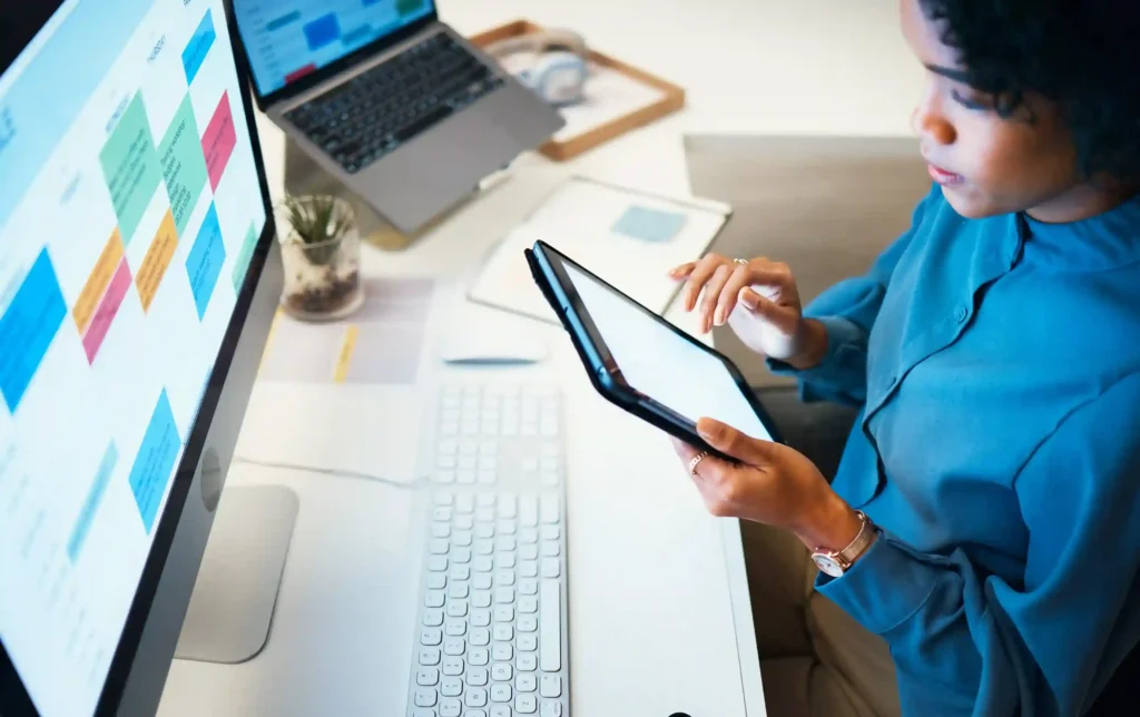 A high-angle shot of a woman at a desk in an office, reviewing a colorful digital schedule on a large desktop monitor while holding and interacting with a tablet.